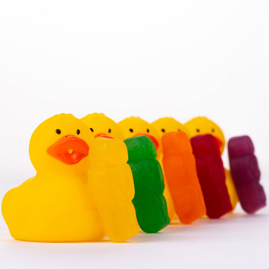 Ducks in a Row 1519 - Close Detail View, vibrant jelly baby figures leaning against a line of yellow rubber duckies, forming a rainbow procession on a white background, fine art photography by Raymond Mather, ART1.