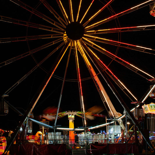 Fairground Carnival Lights 6900065 - Close Detail View, a brightly lit Ferris wheel dominates the night sky, its radiant red, orange, and gold lights forming a dazzling starburst above the motion blur of a spinning ride below. Captured by Raymond Mather, this fine art image evokes the joyful energy and nostalgic charm of a bustling carnival.