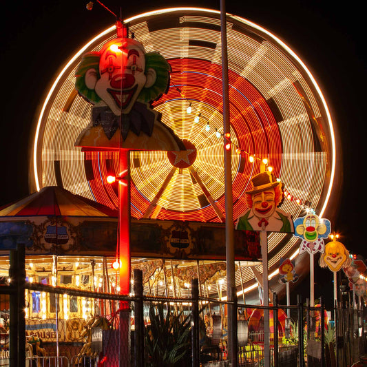 Fairground Carnival Lights 6900090 - Close Detail View, a vivid scene of a spinning Ferris wheel captured in long exposure behind a row of illuminated clown faces and a carousel. Glowing with reds and golds, the image by Raymond Mather evokes the spirited atmosphere of carnival nights, filled with nostalgia, laughter, and motion.