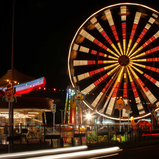 Fairground Carnival Lights 6900103 - Close Detail View, a nighttime carnival scene with a glowing Ferris wheel in spinning motion, colourful string lights, carousel, and clown signs in the foreground. A long exposure adds movement to the entire image, capturing the vibrant pulse of fairground life through Raymond Mather’s lens.