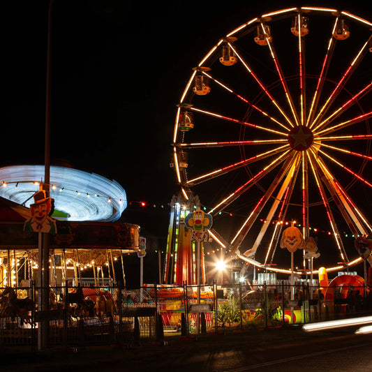 Fairground Carnival Lights 6900108 - Close Detail View, a long exposure nighttime image capturing a glowing Ferris wheel, a spinning carousel in streaks of blue light, and vibrant fairground décor including clown signs and string lights. The photograph by Raymond Mather captures the immersive energy and joy of carnival nights.
