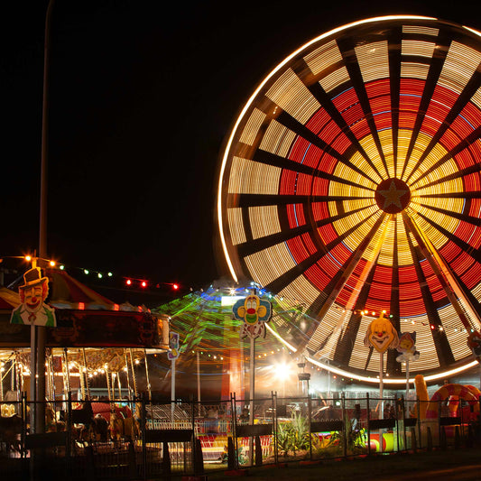 Fairground Carnival Lights 6900129 - Close Detail View, a vibrant Ferris wheel spins in glowing red, yellow, and white light, creating a radiant circular pattern. Below it, a carousel and other rides glow with multicoloured lights and vintage clown decorations. Photographed by Raymond Mather, this image evokes the sensory thrill and nostalgia of carnival nights.