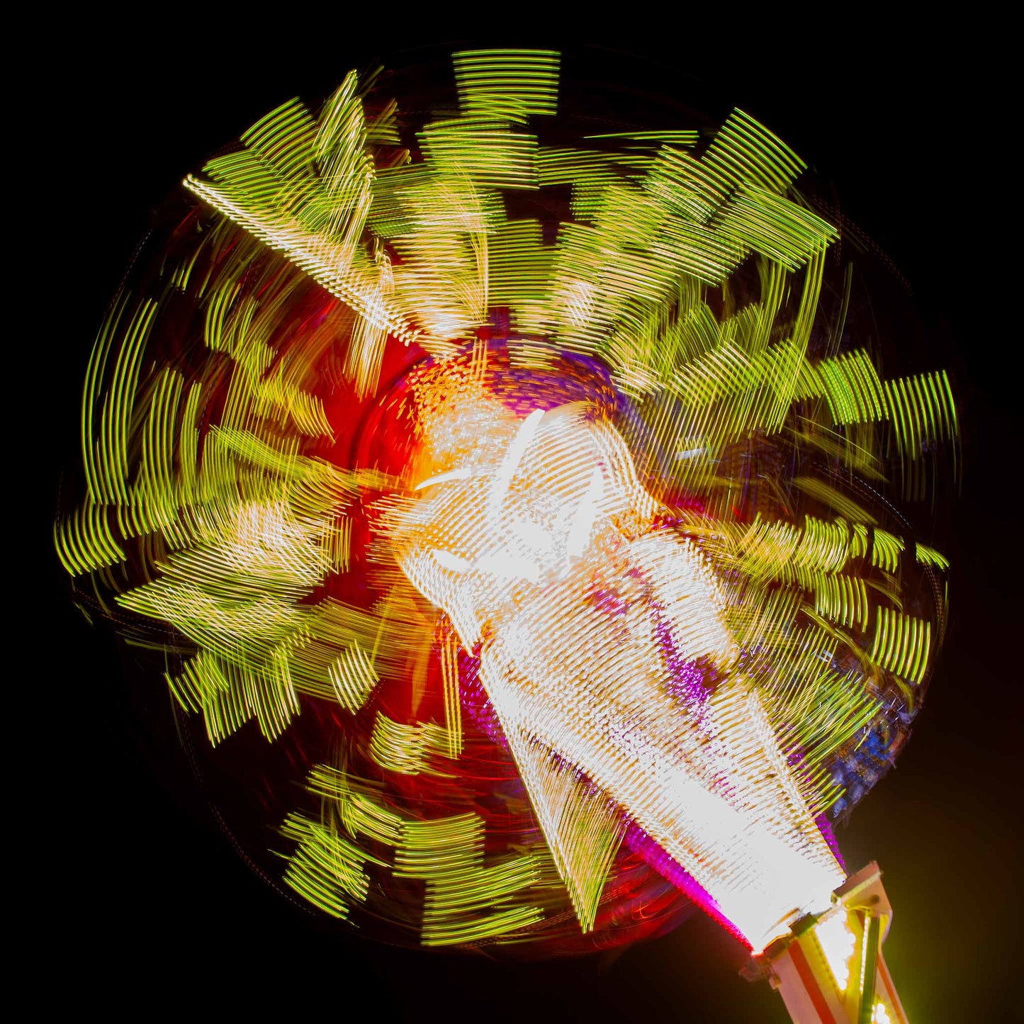 Fairground Carnival Lights 6900132 - 90x90cm, a long exposure photograph of a spinning carnival ride, captured in a dazzling explosion of yellow, red, and white lights radiating from a central axis against a deep black sky. The blurred movement forms abstract, circular patterns, creating a vibrant and kinetic visual effect.