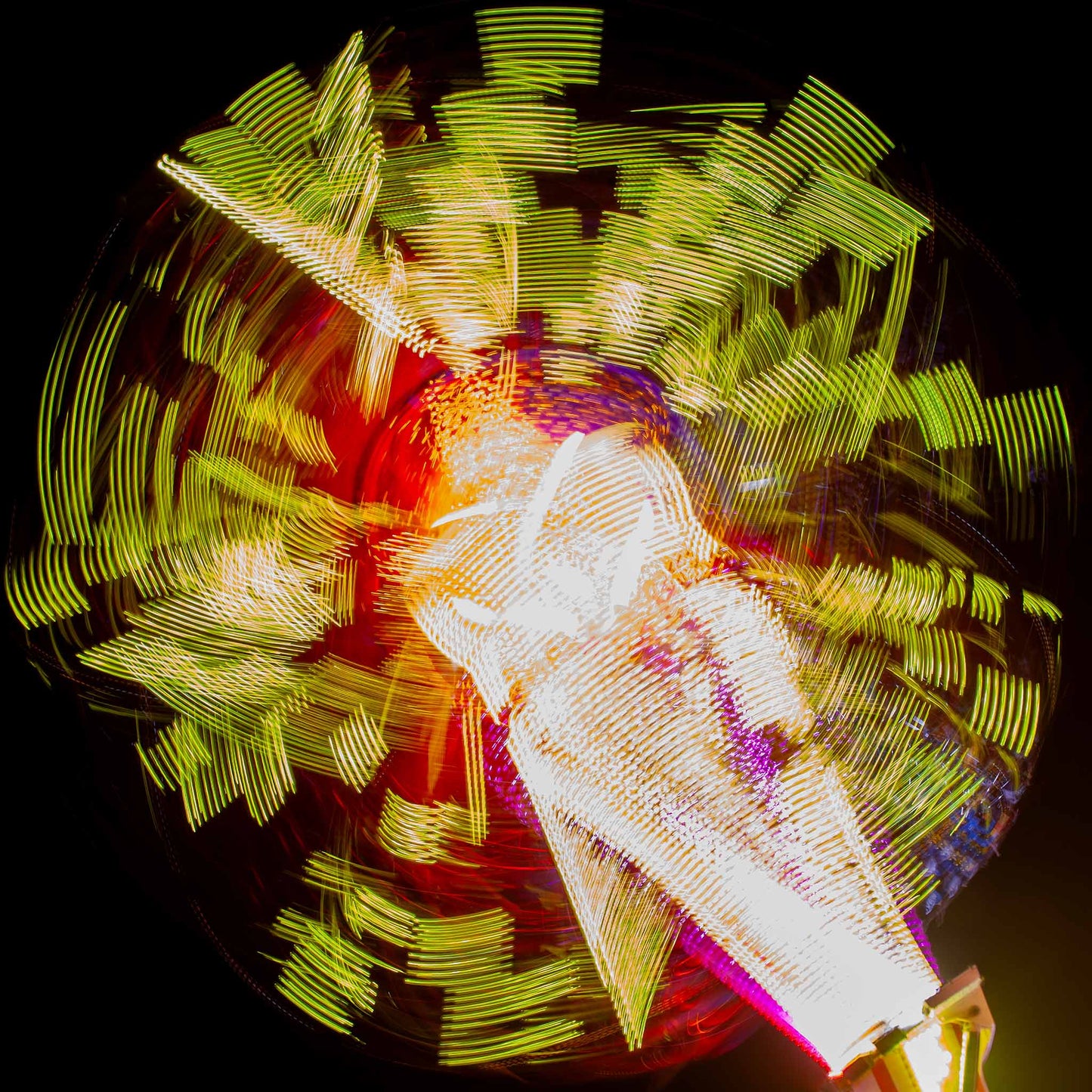Fairground Carnival Lights 6900132 - Close Detail View, a long exposure photograph of a spinning carnival ride, captured in a dazzling explosion of yellow, red, and white lights radiating from a central axis against a deep black sky. The blurred movement forms abstract, circular patterns, creating a vibrant and kinetic visual effect.
