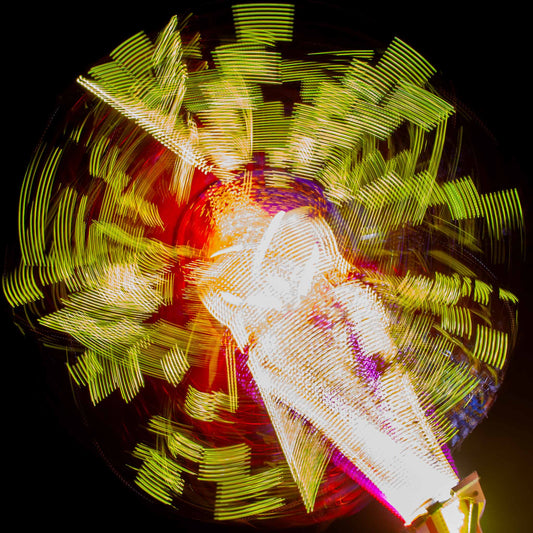 Fairground Carnival Lights 6900132 - Close Detail View, a long exposure photograph of a spinning carnival ride, captured in a dazzling explosion of yellow, red, and white lights radiating from a central axis against a deep black sky. The blurred movement forms abstract, circular patterns, creating a vibrant and kinetic visual effect.