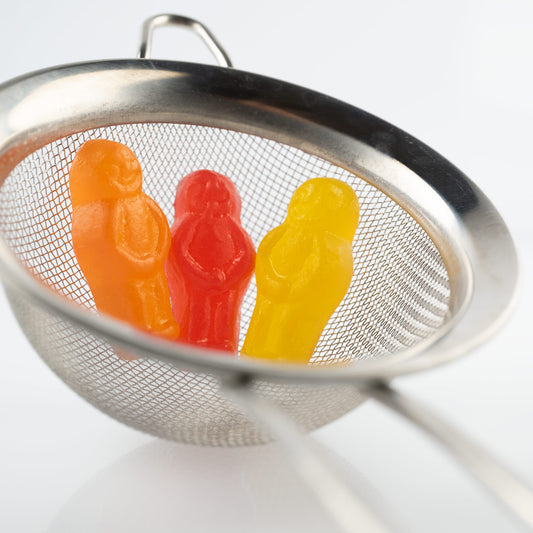 Life’s a Strain 2541v1 - Close Detail View, stylised fine art photograph featuring three vibrant jelly baby figures, orange, red, and yellow, nestled inside a silver tea strainer against a soft white background. A whimsical composition evoking themes of containment, reflection, and resilience, by Raymond Mather, ART1.