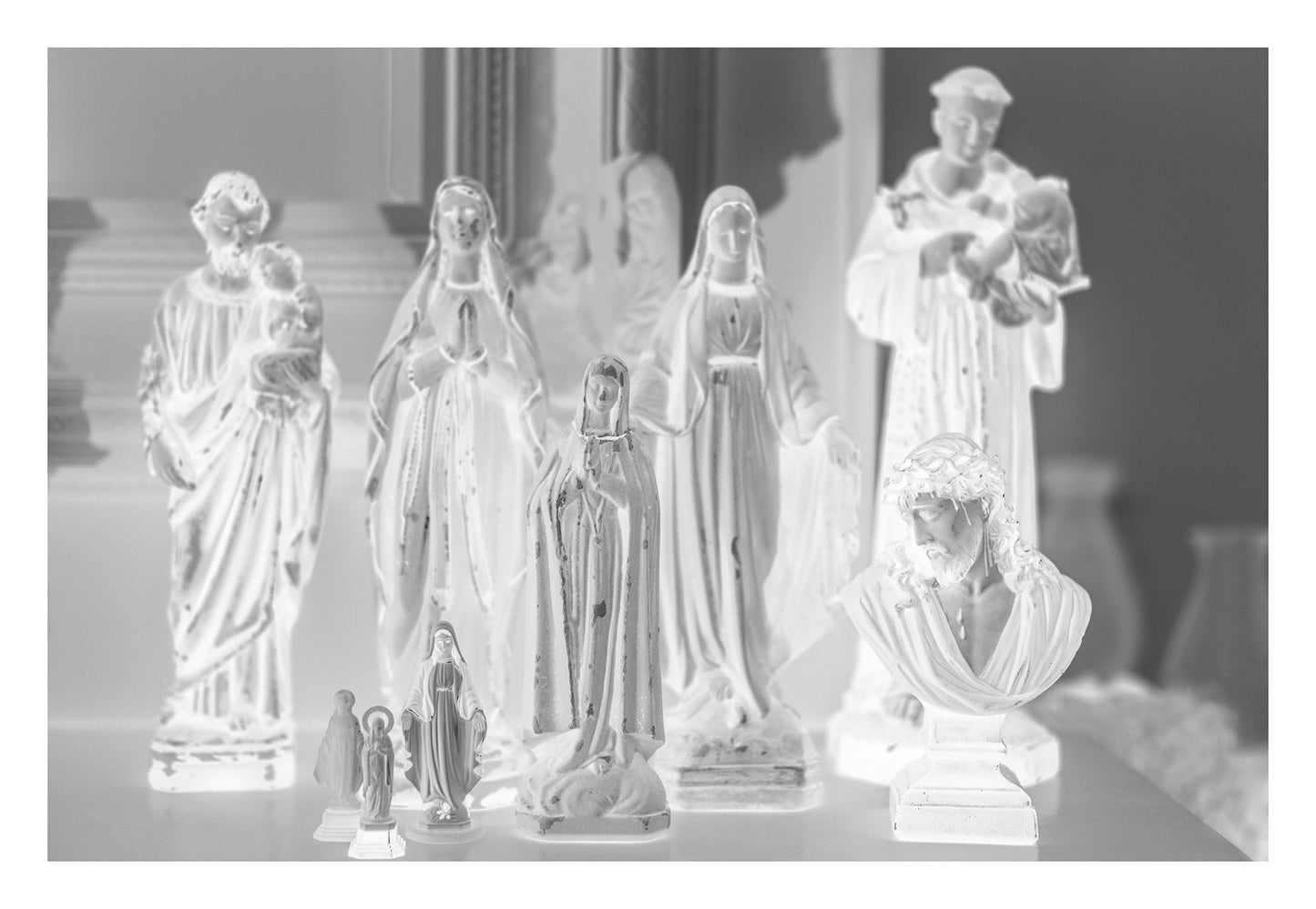 Monochrome Mix - Icons on the Mantel - 183072 - 104x74cm, A monochrome image of religious figurines grouped on a mantel, including various Madonnas, saints, and a bust of Christ, evoking a serene and personal shrine.