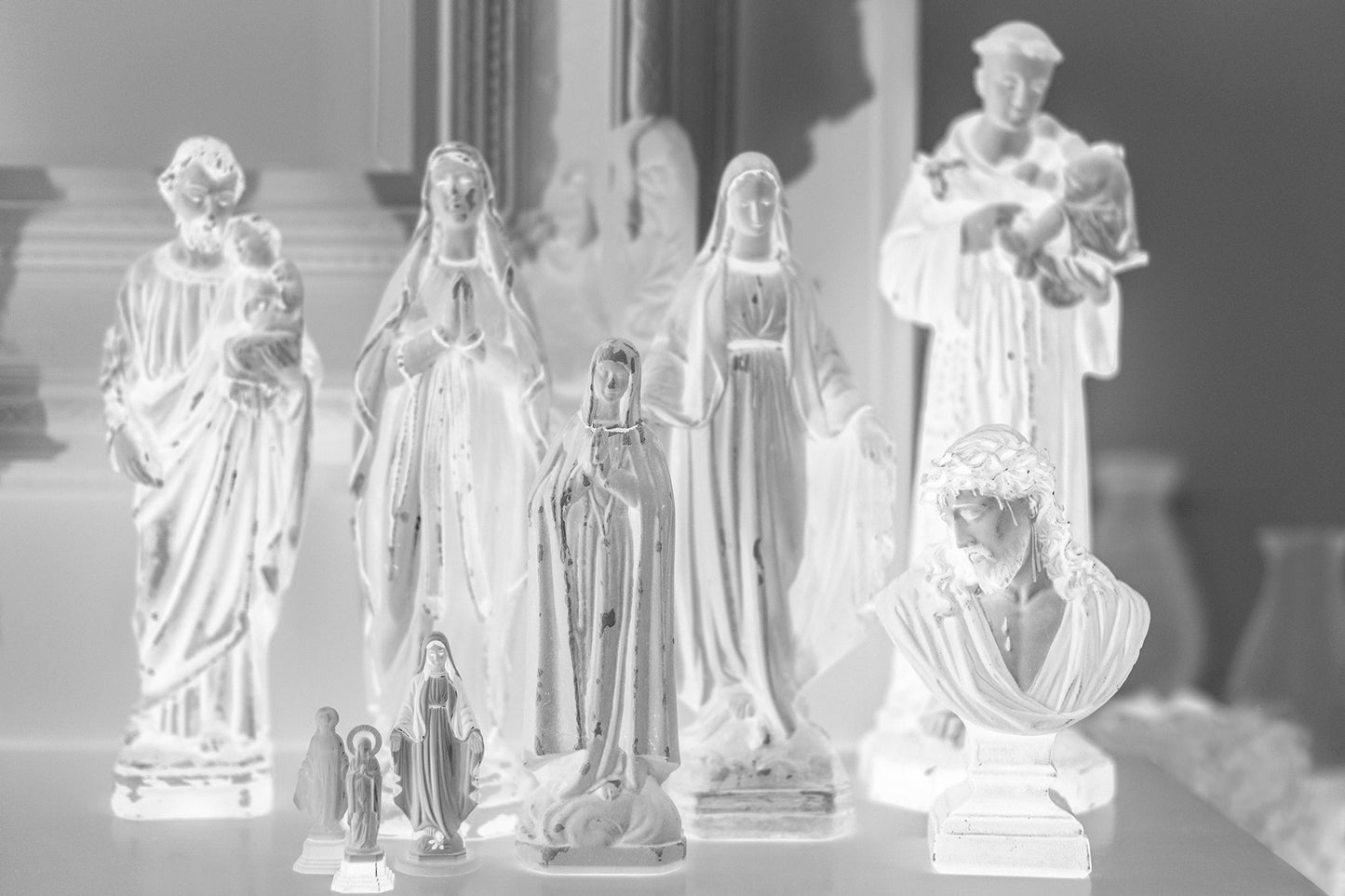 Monochrome Mix - Icons on the Mantel - 183072 - 90x60cm, A monochrome image of religious figurines grouped on a mantel, including various Madonnas, saints, and a bust of Christ, evoking a serene and personal shrine.