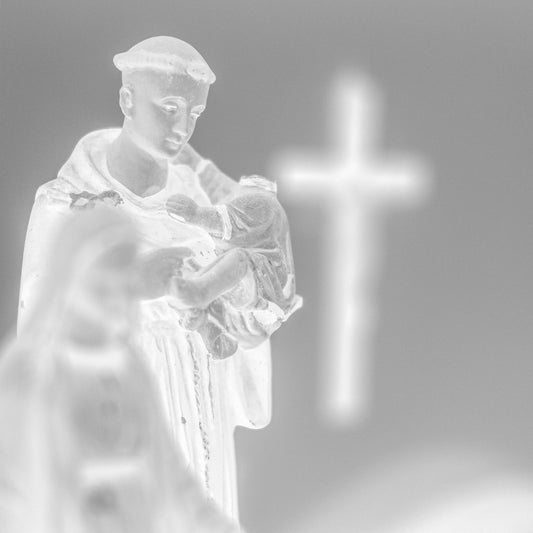 Monochrome Mix - Icons on the Mantel - 183102 - Close Detail View, Black and white image of a Saint Anthony statue holding baby Jesus, with a glowing cross in the blurred background, evoking spiritual reflection and reverent stillness.