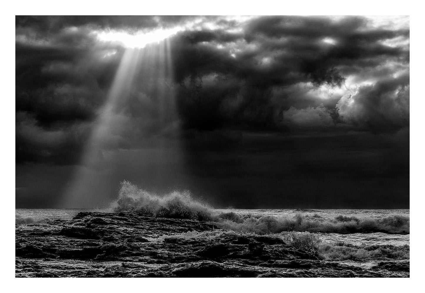 Monochrome Mix - Valla Beach - Cloud Burst - 171683 - 104x74cm, Black and white photograph of stormy ocean waves crashing on rocks beneath dramatic cloud cover, with beams of sunlight piercing through the clouds over Valla Beach.