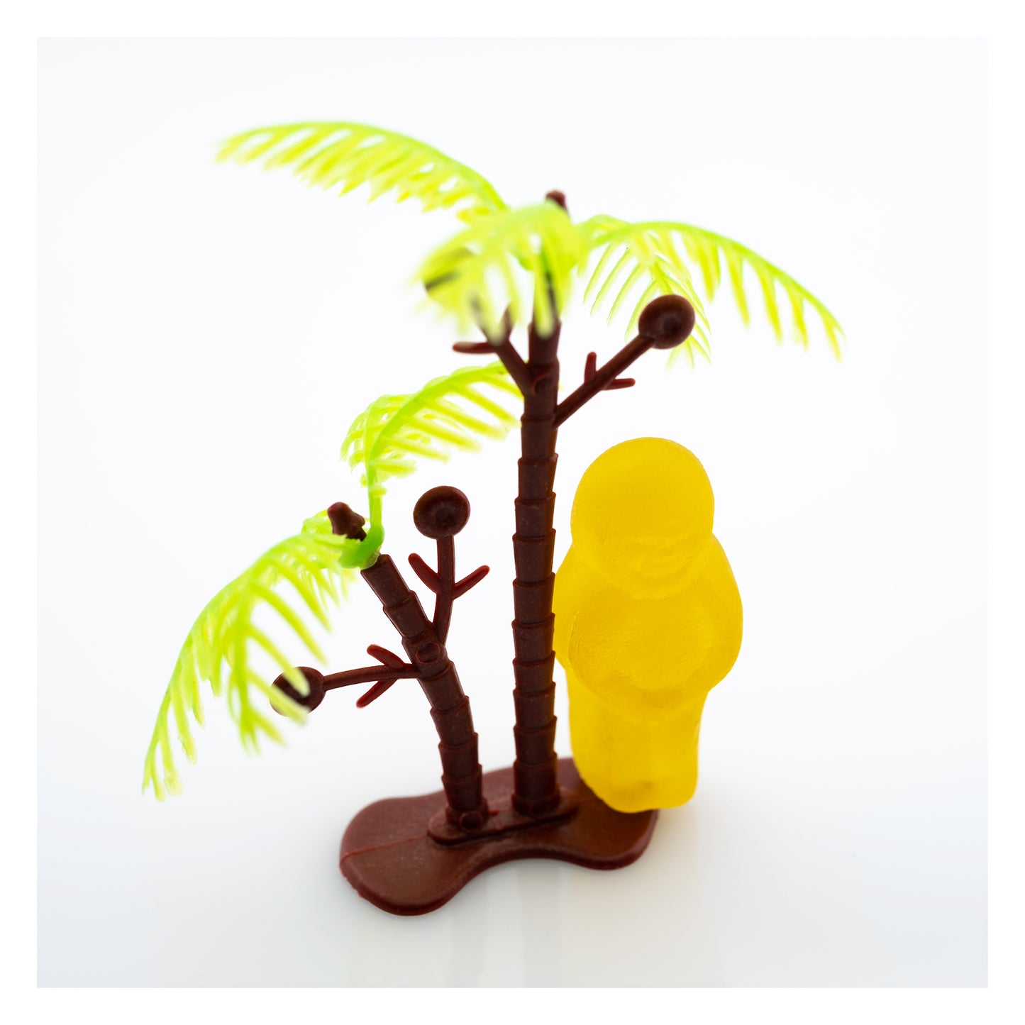 No Man Is an Island 3000v2 - 104x104cm, fine art conceptual photograph by Raymond Mather featuring a solitary yellow jelly baby figure standing beside a miniature palm tree on a pristine white background, symbolising isolation and whimsy through playful visual metaphor.