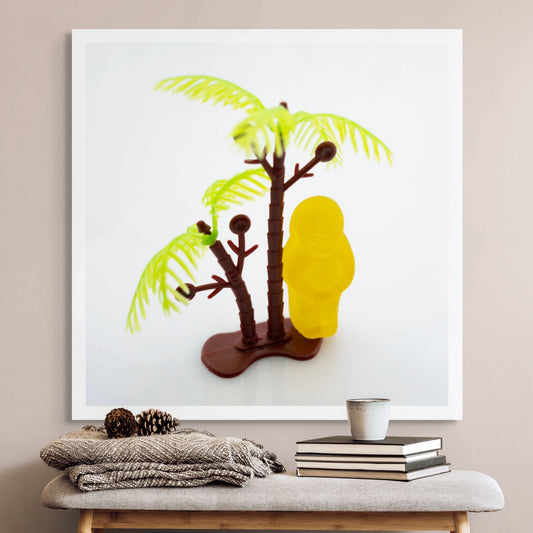 No Man Is an Island 3000v2 - Room View, fine art conceptual photograph by Raymond Mather featuring a solitary yellow jelly baby figure standing beside a miniature palm tree on a pristine white background, symbolising isolation and whimsy through playful visual metaphor.