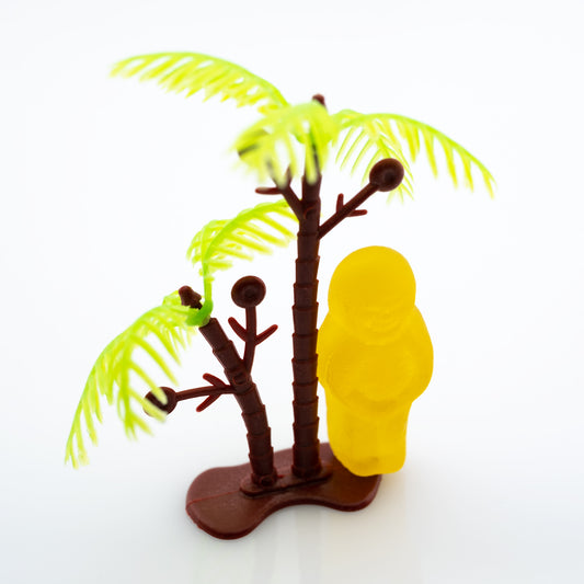 No Man Is an Island 3000v2 - Close Detail View, fine art conceptual photograph by Raymond Mather featuring a solitary yellow jelly baby figure standing beside a miniature palm tree on a pristine white background, symbolising isolation and whimsy through playful visual metaphor.
