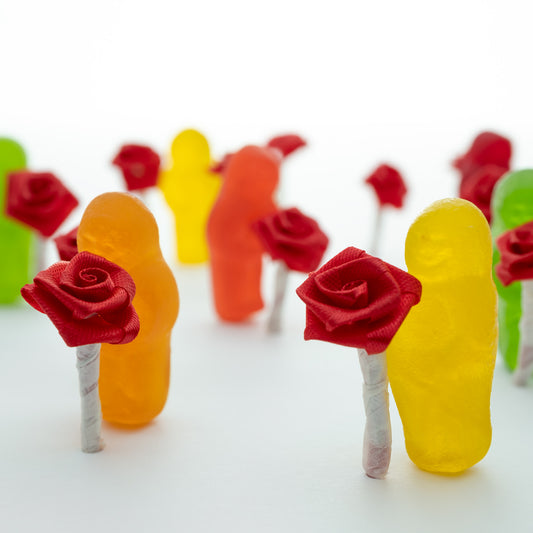 Stop and Smell the Roses 2836v1 - Close Detail View, playful fine art photograph by Raymond Mather featuring colourful jelly baby figures holding miniature red ribbon roses, arranged in a dreamlike procession on a soft white backdrop, evoking mindfulness and delight.