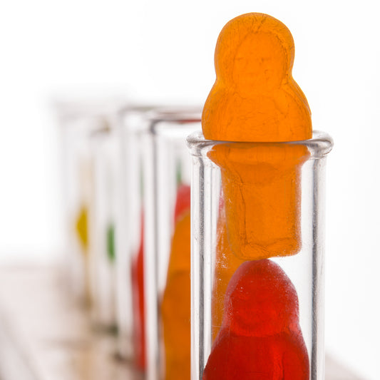 Test Tube Babies 20101 - Close Detail View, fine art photograph by Raymond Mather featuring brightly coloured jelly baby figures inside clear glass test tubes arranged in a wooden rack, a playful yet thought provoking pop art tableau exploring identity, science, and the absurdity of cliché.