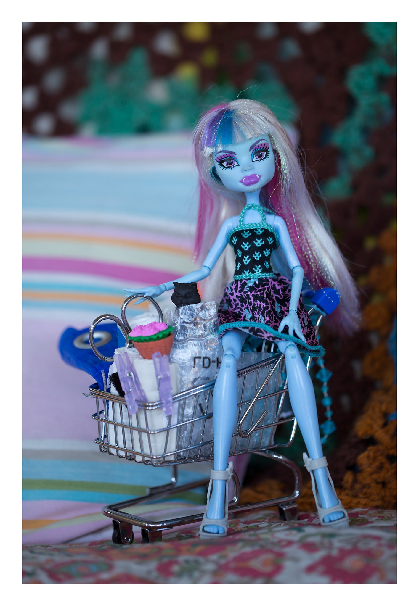 Trolley Dolly - Series No.1: 197147 - 74x104cm, a blue skinned fashion doll with pastel hair sits inside a miniature shopping trolley filled with handmade props including a cupcake, bag, and clipped paper rolls. Set against a striped and crocheted background, the image explores themes of homelessness, childhood, and resilience.