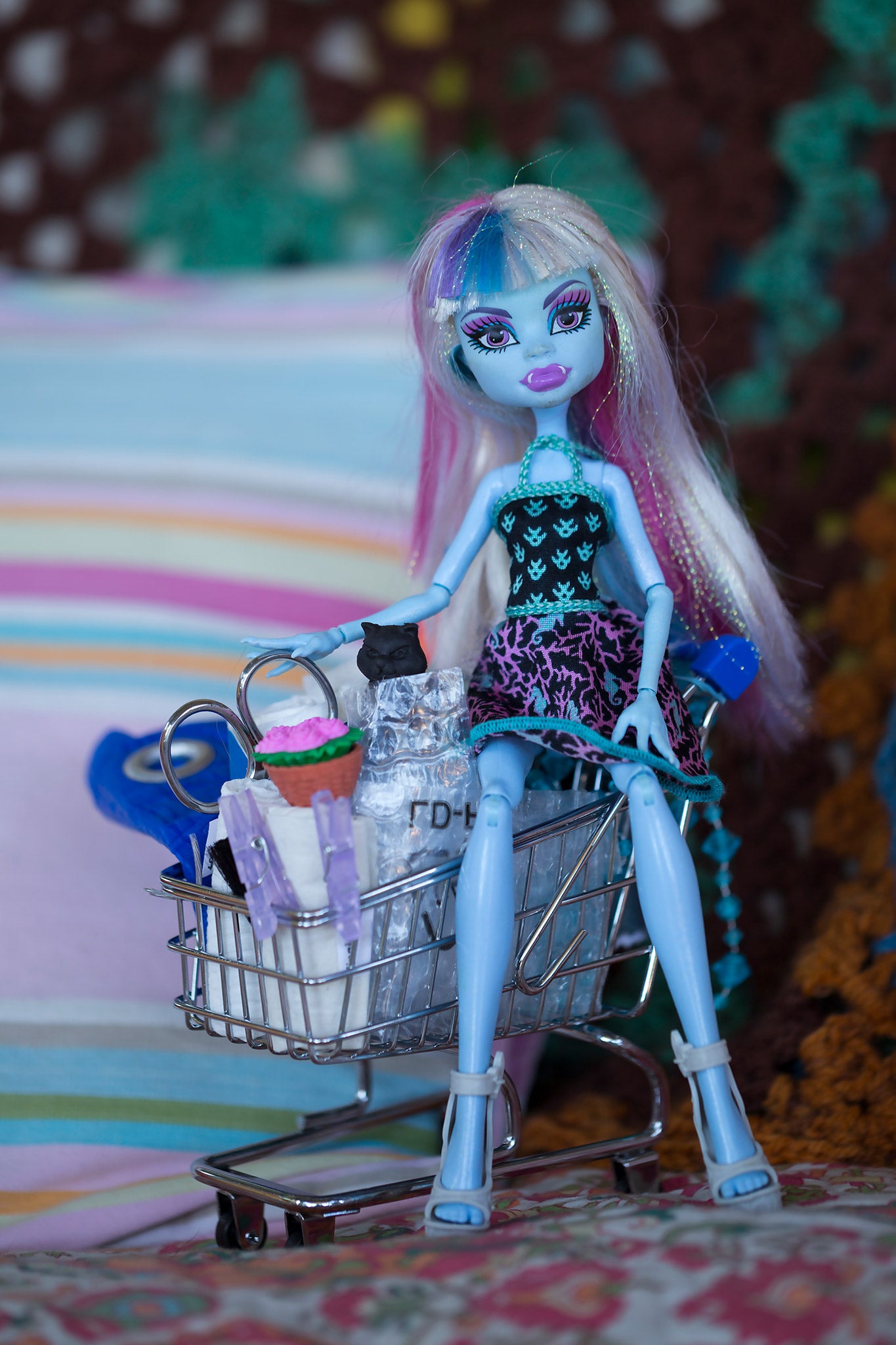 Trolley Dolly - Series No.1: 197147 - 60x90cm, a blue skinned fashion doll with pastel hair sits inside a miniature shopping trolley filled with handmade props including a cupcake, bag, and clipped paper rolls. Set against a striped and crocheted background, the image explores themes of homelessness, childhood, and resilience.