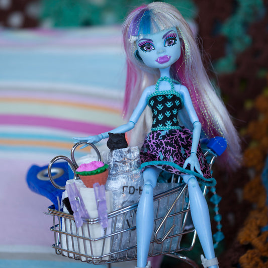Trolley Dolly - Series No.1: 197147 - Close Detail View, a blue skinned fashion doll with pastel hair sits inside a miniature shopping trolley filled with handmade props including a cupcake, bag, and clipped paper rolls. Set against a striped and crocheted background, the image explores themes of homelessness, childhood, and resilience.
