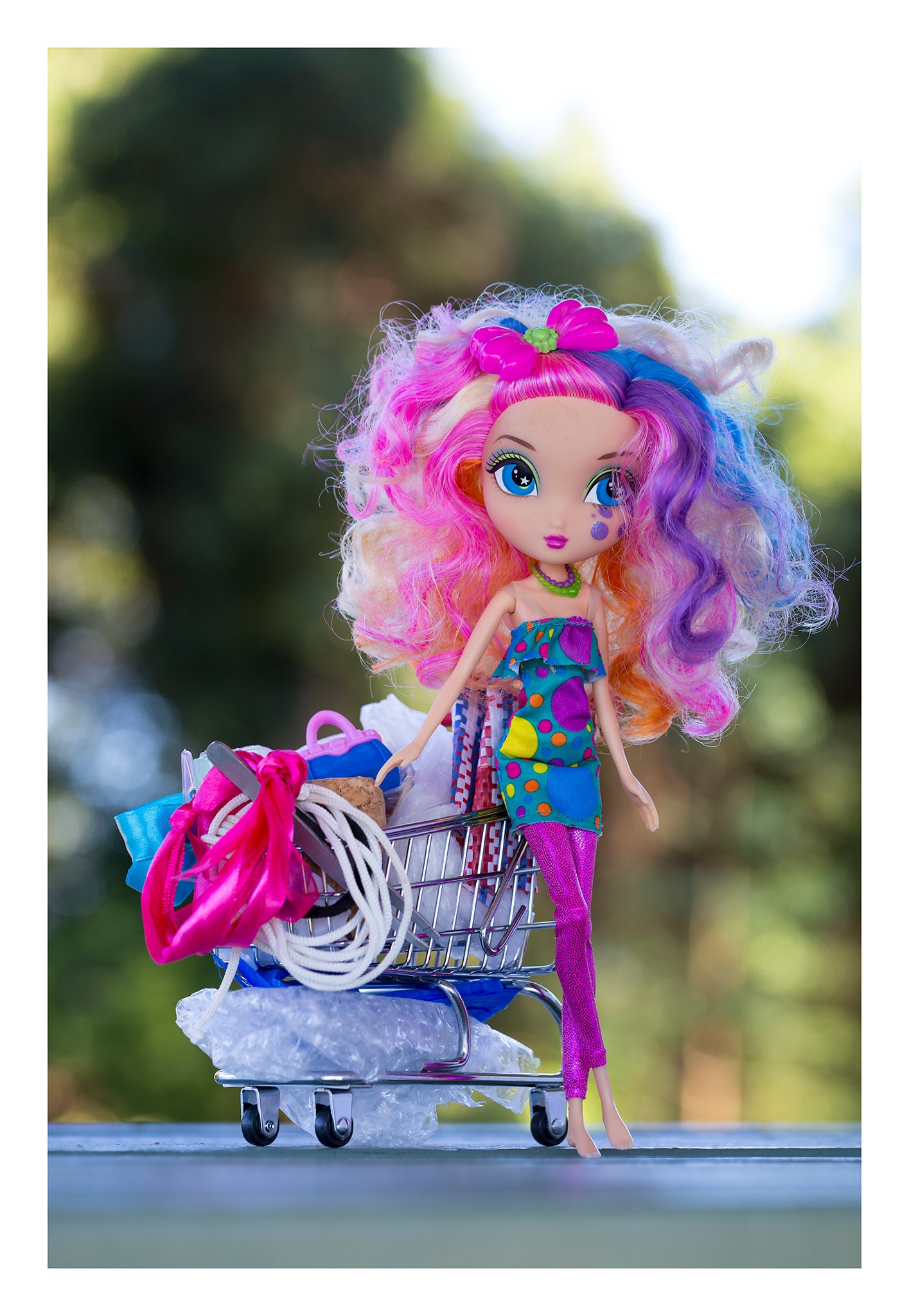 Trolley Dolly - Series No.1: 197260 - 74x104cm, a colourful fashion doll with curly rainbow hair and a tear on her cheek stands beside an overflowing miniature shopping trolley packed with symbolic items like ribbons, fabric, and string. Set against a soft outdoor backdrop, the image explores displacement, resilience, and hidden stories.