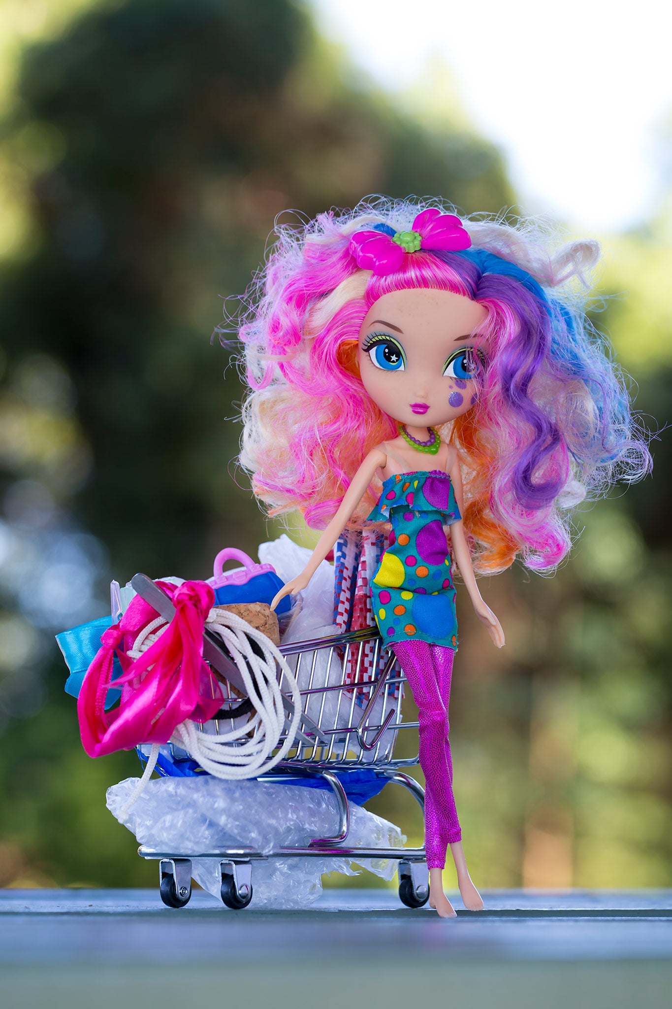 Trolley Dolly - Series No.1: 197260 - 60x90cm, a colourful fashion doll with curly rainbow hair and a tear on her cheek stands beside an overflowing miniature shopping trolley packed with symbolic items like ribbons, fabric, and string. Set against a soft outdoor backdrop, the image explores displacement, resilience, and hidden stories.