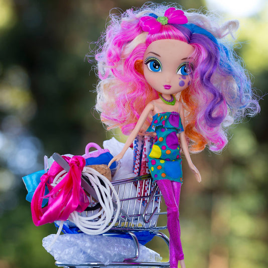 Trolley Dolly - Series No.1: 197260 - Close Detail View, a colourful fashion doll with curly rainbow hair and a tear on her cheek stands beside an overflowing miniature shopping trolley packed with symbolic items like ribbons, fabric, and string. Set against a soft outdoor backdrop, the image explores displacement, resilience, and hidden stories.