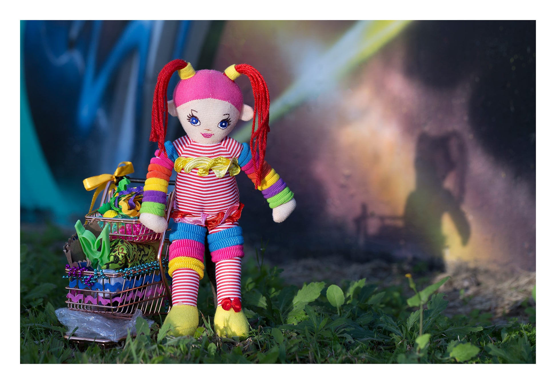 Trolley Dolly - Series No.1: 197291 - 104x74cm, a colourful handmade cloth doll with red pigtails stands beside a mini trolley filled with ribbons, trinkets, and found objects. Her shadow is cast onto a galaxy style mural wall in the background. The image explores homelessness, identity, and play through symbolic layering.