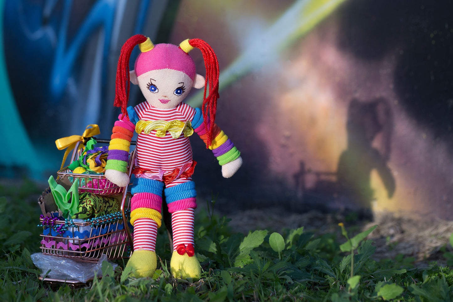Trolley Dolly - Series No.1: 197291 - 90x60cm, a colourful handmade cloth doll with red pigtails stands beside a mini trolley filled with ribbons, trinkets, and found objects. Her shadow is cast onto a galaxy style mural wall in the background. The image explores homelessness, identity, and play through symbolic layering.