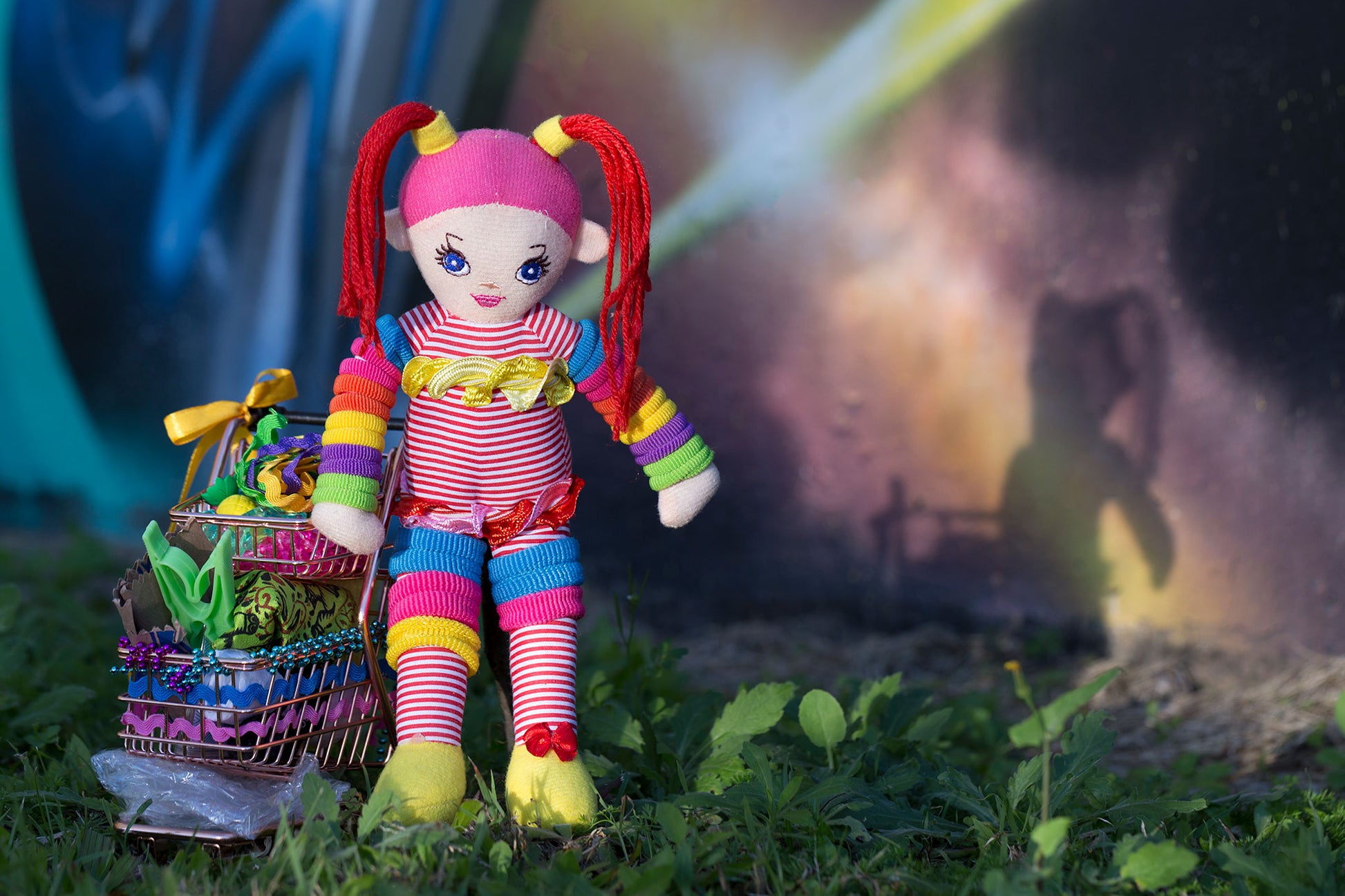 Trolley Dolly - Series No.1: 197291 - 90x60cm, a colourful handmade cloth doll with red pigtails stands beside a mini trolley filled with ribbons, trinkets, and found objects. Her shadow is cast onto a galaxy style mural wall in the background. The image explores homelessness, identity, and play through symbolic layering.