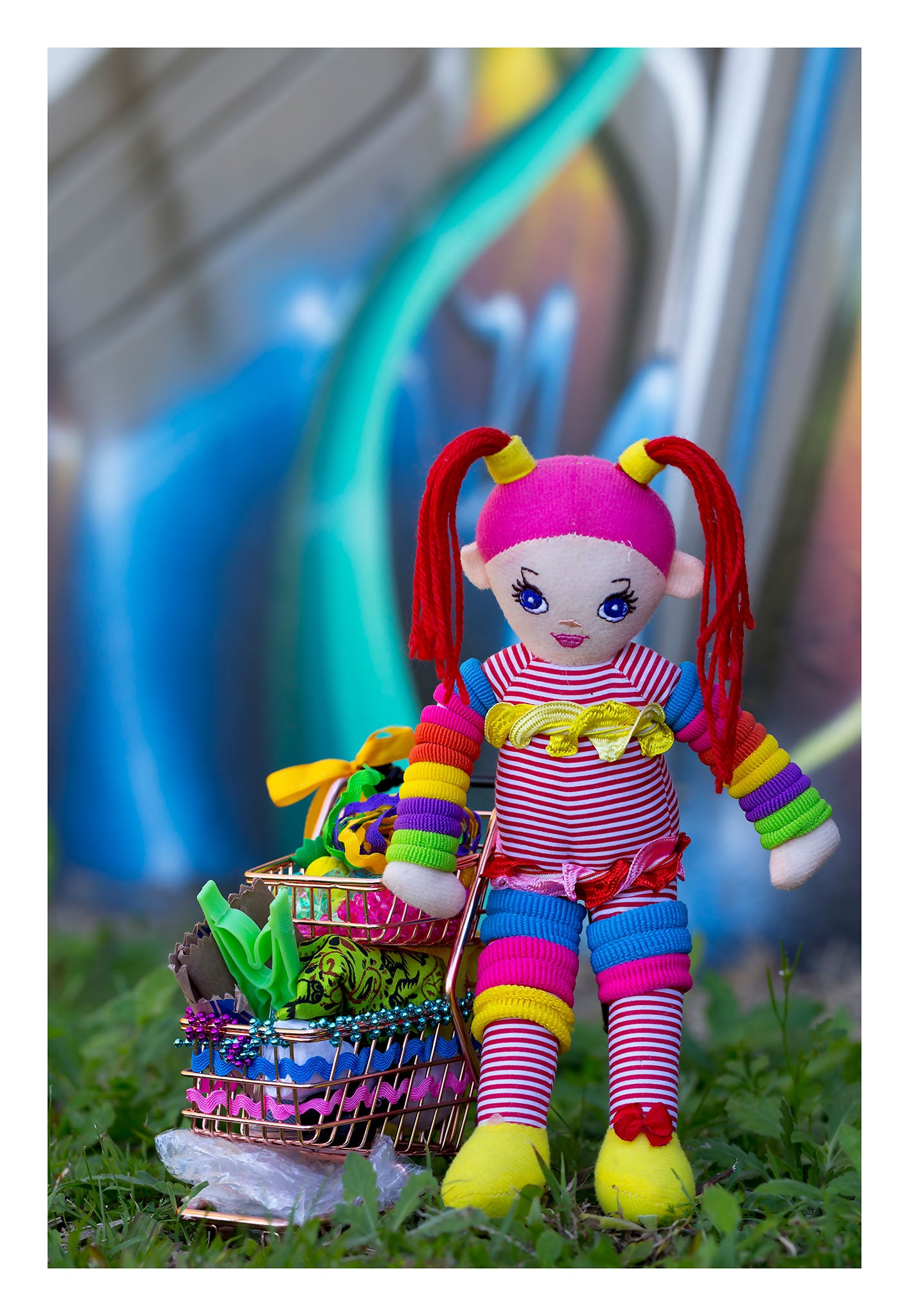 Trolley Dolly - Series No.1: 197302 - 74x104cm, a handmade rainbow coloured rag doll stands beside a mini shopping trolley filled with ribbons, beads, fabric, and found objects. Set on grass against a vivid urban graffiti wall, the image explores homelessness, survival, and innocence.