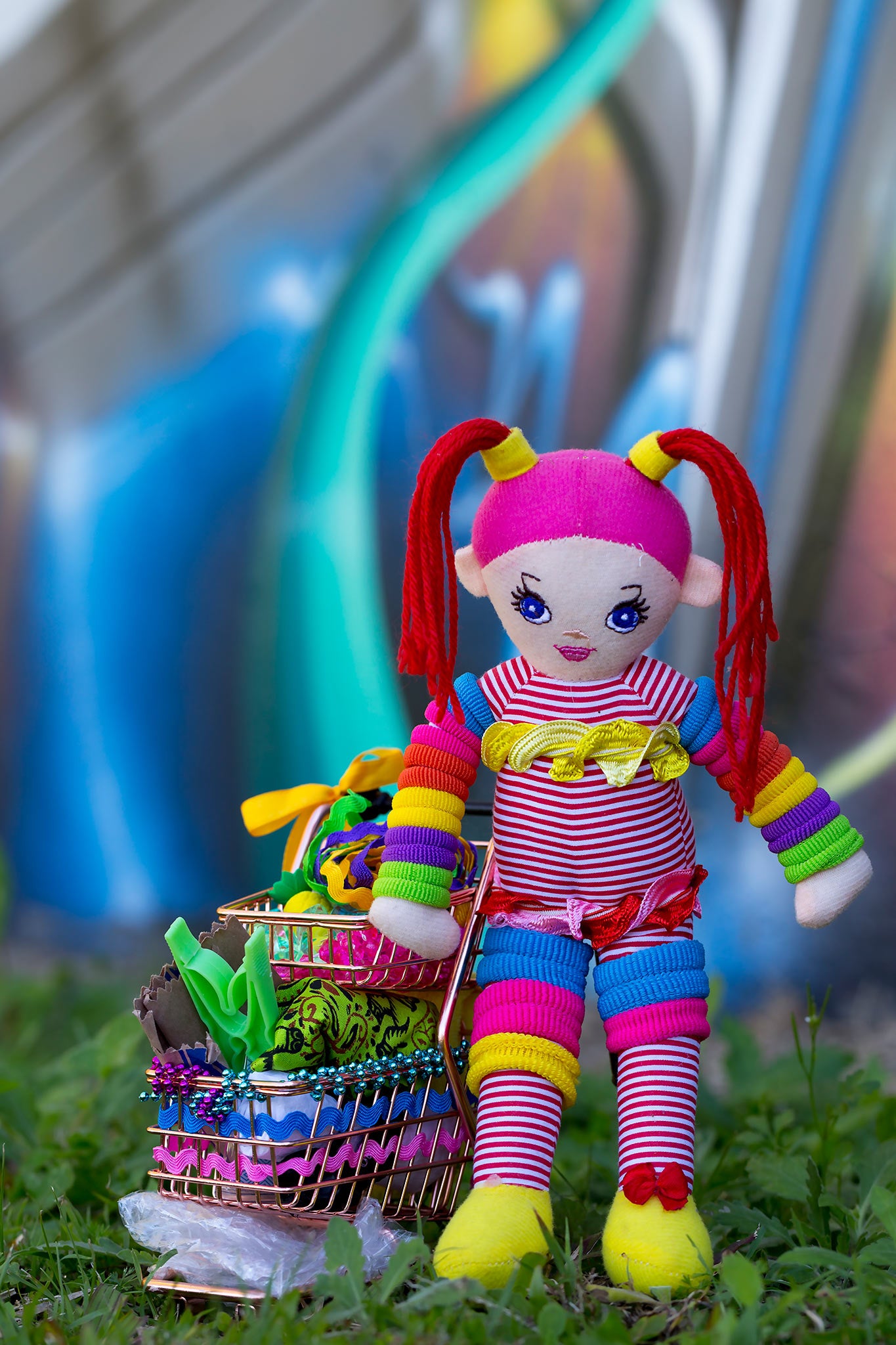 Trolley Dolly - Series No.1: 197302 - 60x90cm, a handmade rainbow coloured rag doll stands beside a mini shopping trolley filled with ribbons, beads, fabric, and found objects. Set on grass against a vivid urban graffiti wall, the image explores homelessness, survival, and innocence.