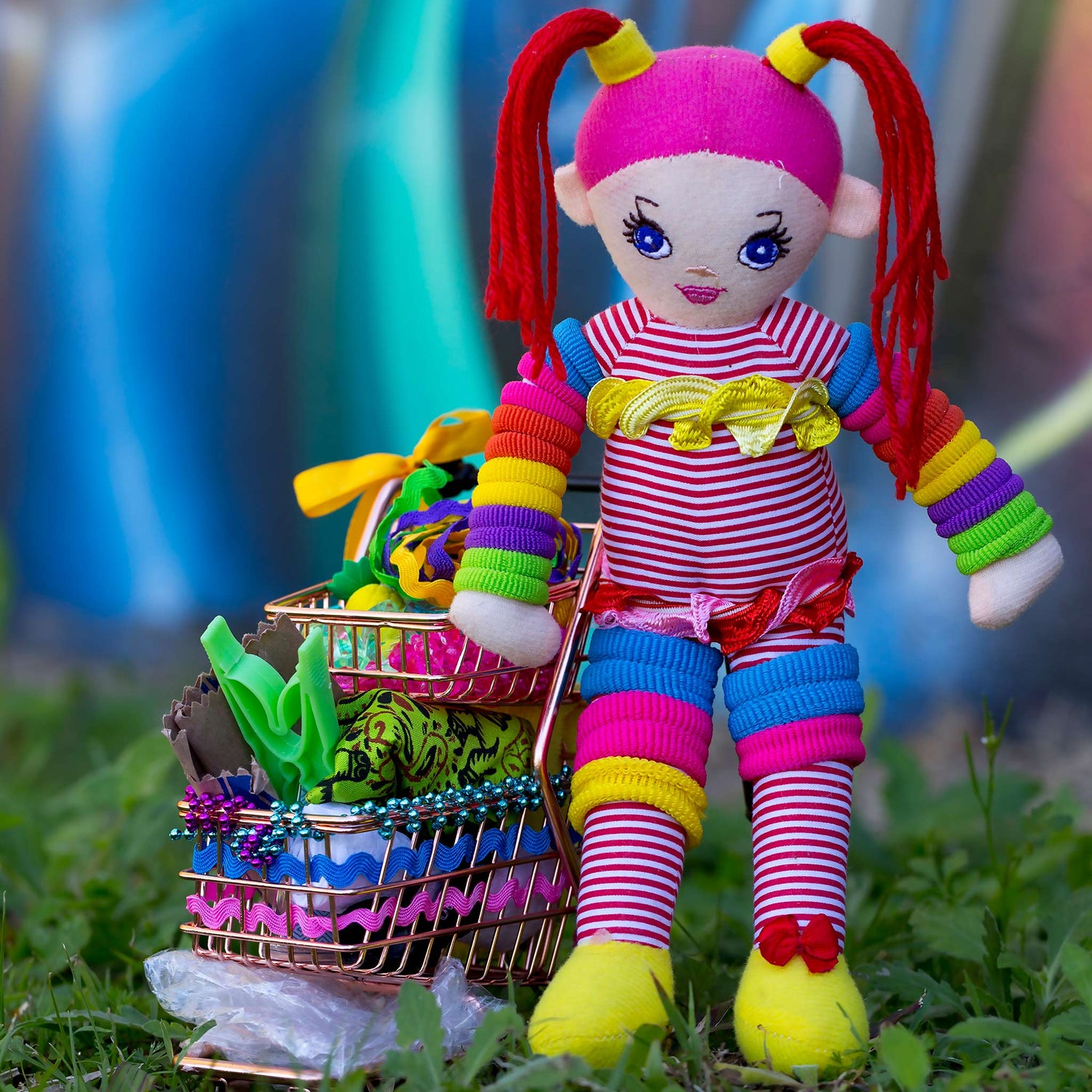 Trolley Dolly - Series No.1: 197302 - Close Detail View, a handmade rainbow coloured rag doll stands beside a mini shopping trolley filled with ribbons, beads, fabric, and found objects. Set on grass against a vivid urban graffiti wall, the image explores homelessness, survival, and innocence.