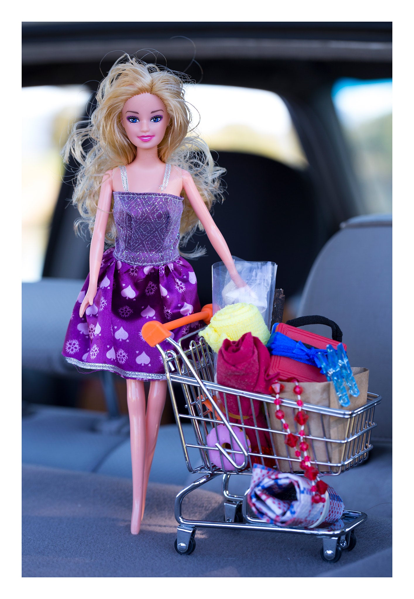 Trolley Dolly - Series No.1: 197392 - 74x104cm, a blonde fashion doll in a purple dress stands beside a miniature trolley filled with fabric, clips, beads, and essentials, photographed inside a car. The image explores hidden homelessness and the feminine burden of appearing composed amid survival.