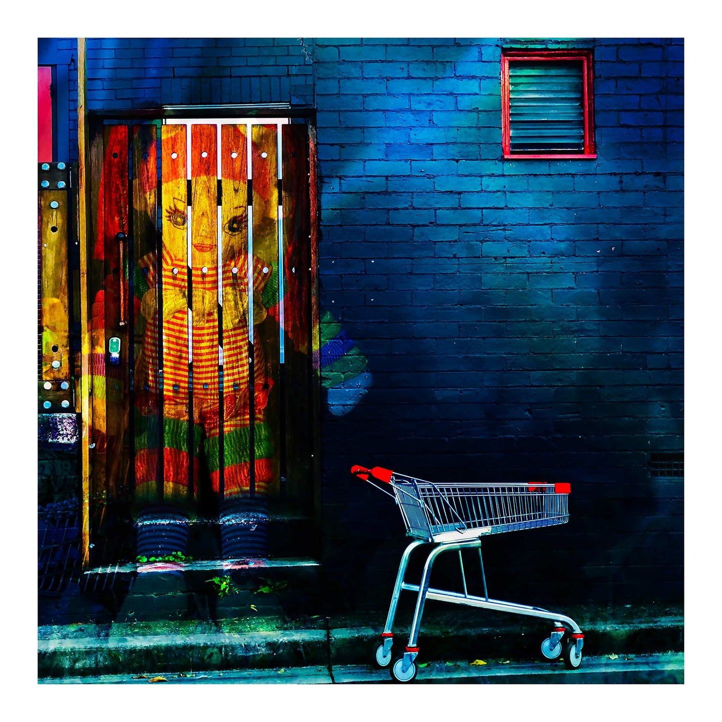Trolley Dolly - Series No.2: 19E2255v13 - 74x74cm, a colourful knitted doll appears embedded in a wooden door, her red, green, and striped outfit vivid against a moody blue brick wall. An empty red handled shopping trolley sits at the kerbside. The composition symbolises childhood, homelessness, and the silent resilience of unseen lives.