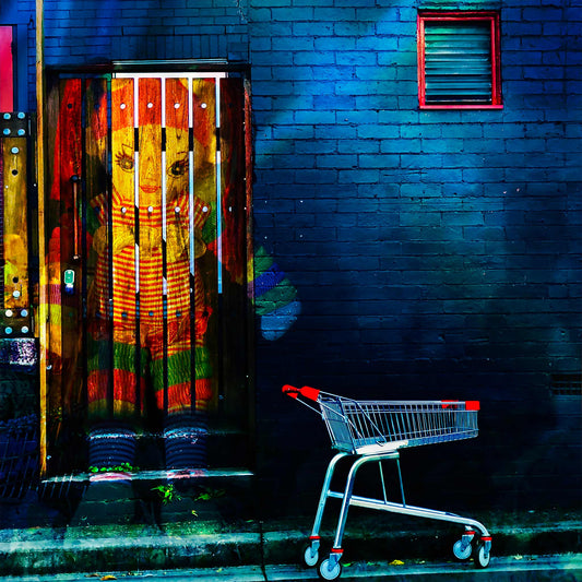 Trolley Dolly - Series No.2: 19E2255v13 - Close Detail View, a colourful knitted doll appears embedded in a wooden door, her red, green, and striped outfit vivid against a moody blue brick wall. An empty red handled shopping trolley sits at the kerbside. The composition symbolises childhood, homelessness, and the silent resilience of unseen lives.