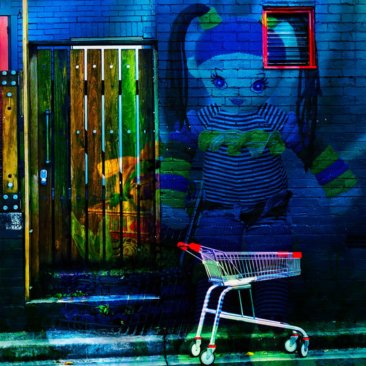 Trolley Dolly - Series No.2: 19E2255v14 - Close Detail View, a knitted doll with striped clothing and ponytails appears against a dark blue brick wall. A red-handled shopping trolley sits below, and a wooden door to the left glows with market textures and shadow. The surreal image explores homelessness and emotional vulnerability.