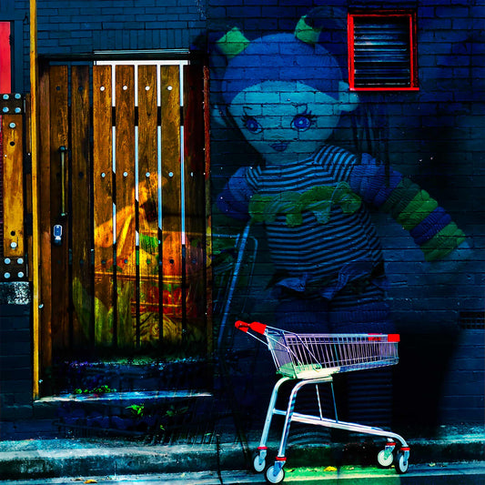 Trolley Dolly - Series No.2: 9255v3 - Close Detail View, a surreal urban street scene featuring a large doll overlaid against a dark brick wall, her striped outfit and outstretched arms glowing in soft hues. In the foreground, a shopping trolley with red handles is parked on the footpath. A textured wooden door and a red trimmed vent hint at hidden stories. The layered composition conveys a dreamlike tension between innocence and abandonment, reflecting on homelessness and social neglect.