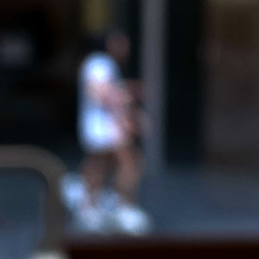 Urban Commute - Series No.1: 0979v1 - Close Detail View, a soft focus photograph of two city walkers side by side, dressed in light tones with glowing white shoes, set against a deep urban backdrop. A gesture of shared movement and quiet rhythm. Limited edition fine art print by Raymond Mather.