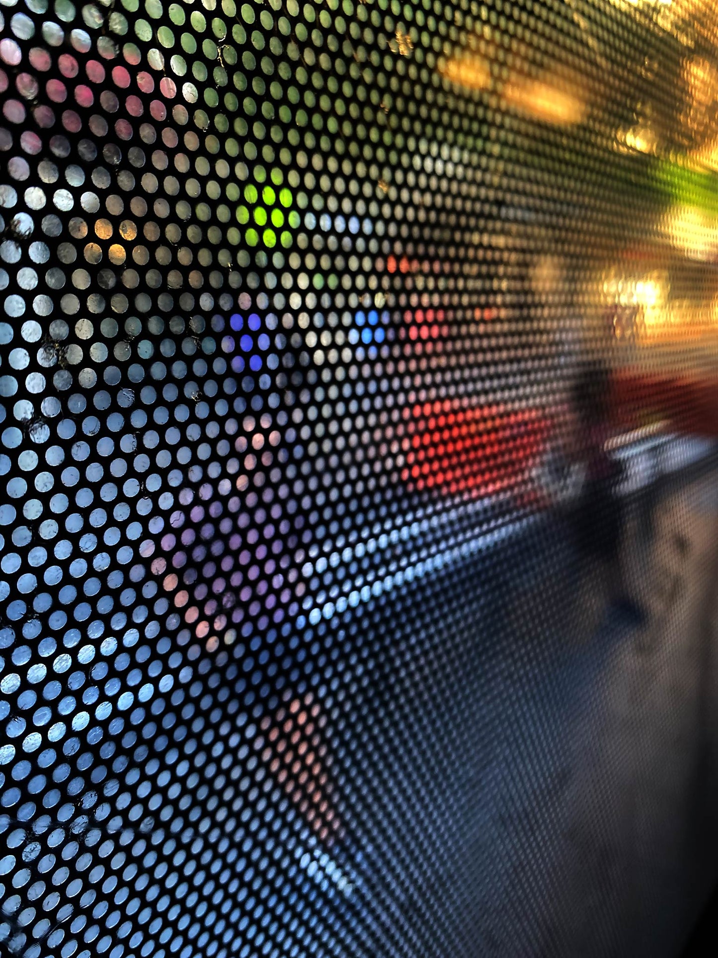 Urban Commute - Series No.2: 0785 - 67.5x90cm, fine art photograph by Raymond Mather featuring a blurred pedestrian viewed through a perforated window screen. Vibrant colours of red, purple, and green shimmer across the gridded surface, creating a dynamic abstract portrait of city life in motion.