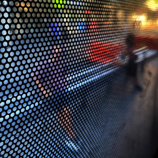 Urban Commute - Series No.2: 0785 - Close Detail View, fine art photograph by Raymond Mather featuring a blurred pedestrian viewed through a perforated window screen. Vibrant colours of red, purple, and green shimmer across the gridded surface, creating a dynamic abstract portrait of city life in motion.