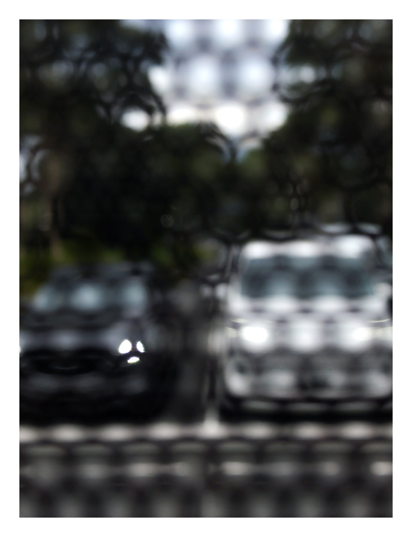 Urban Commute - Series No.2: 8468(1)v1 - 81.5x104cm, fine art photograph by Raymond Mather featuring a soft-focus image of two parked cars, one dark, one white, viewed through a textured, patterned screen. The effect blurs details and creates a moody, abstract urban vignette rich in contrast and stillness.