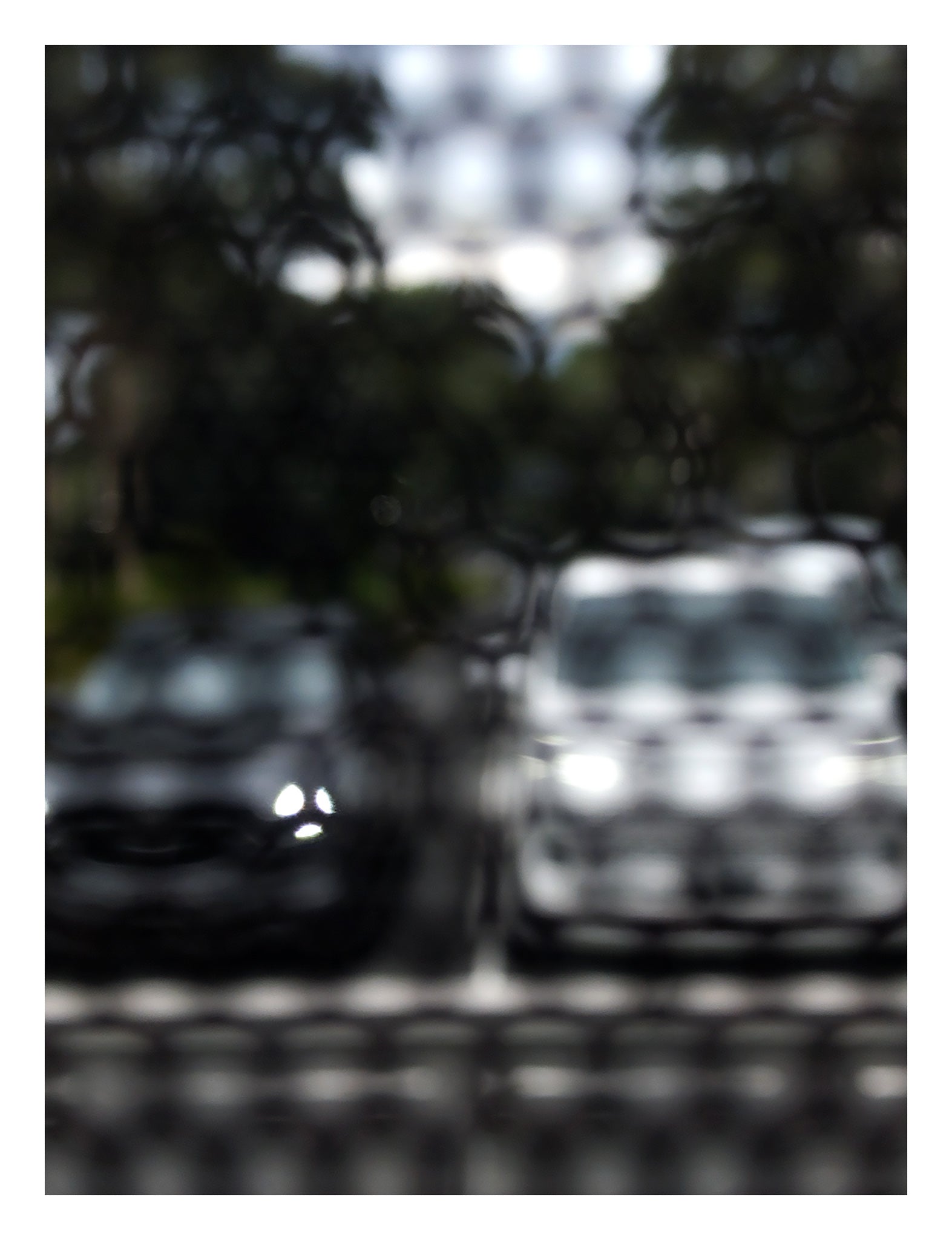 Urban Commute - Series No.2: 8468(1)v1 - 81.5x104cm, fine art photograph by Raymond Mather featuring a soft-focus image of two parked cars, one dark, one white, viewed through a textured, patterned screen. The effect blurs details and creates a moody, abstract urban vignette rich in contrast and stillness.