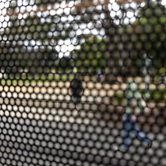 Urban Commute - Series No.2: 8475v1 - Close Detail View, fine art photograph by Raymond Mather featuring two pedestrians viewed through a dark circular grid. One figure in blue jeans moves mid-stride, while the other recedes into a background of trees and dappled light. A soft-focus urban composition exploring movement, texture, and visual rhythm.