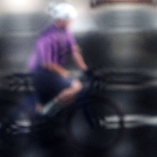 Urban Commute, Series No.2: 8621(1)v1 - Close Detail View, fine art photograph by Raymond Mather featuring a blurred cyclist in purple shirt and white helmet, riding through a moody, reflective cityscape. The image is softly distorted, blending motion with atmosphere in a dreamlike urban abstraction.