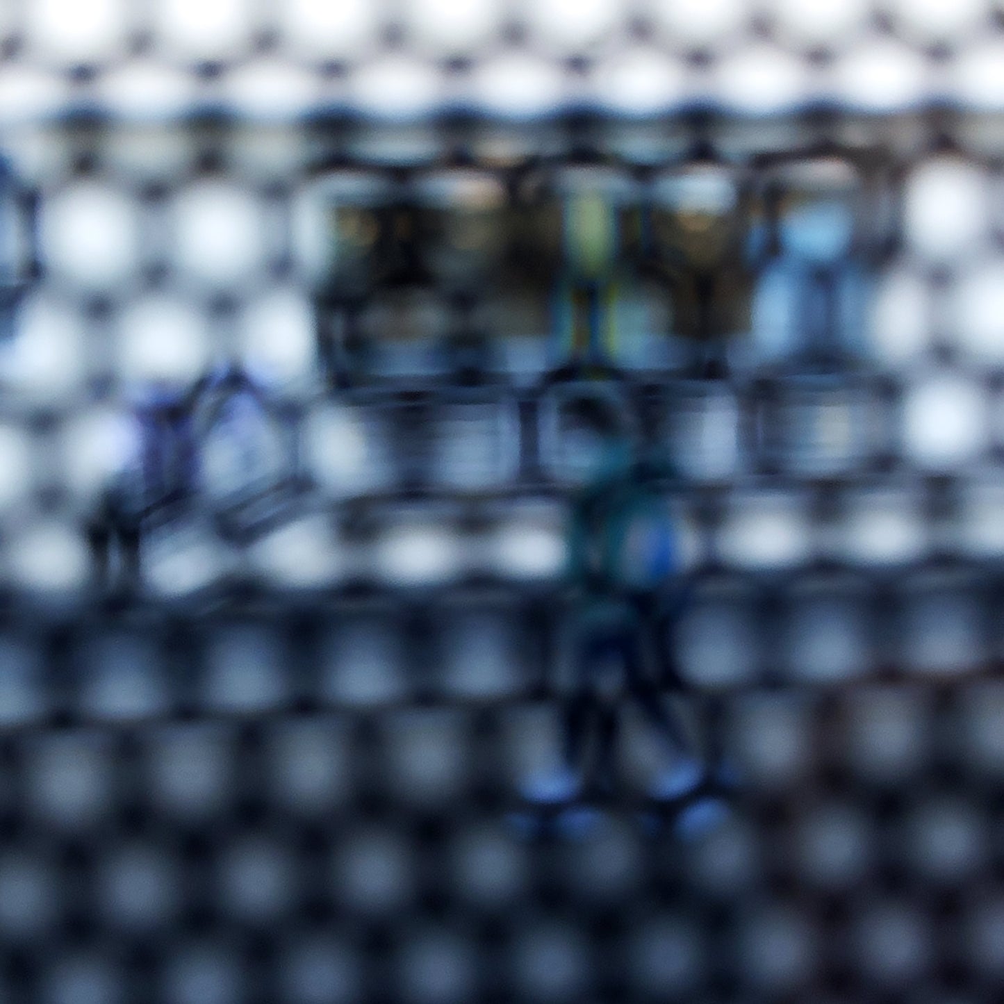 Urban Commute - Series No.2: 9150 - Close Detail View, fine art photograph by Raymond Mather showing a walking figure in blue, captured through a dense circular grid. A blurred train forms the backdrop, creating a quiet, geometric abstraction of city life in motion.