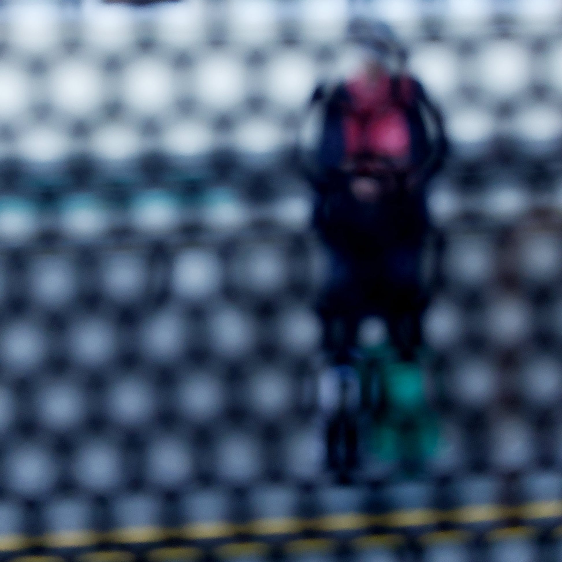 Urban Commute - Series No.2: 9153 - Close Detail View, fine art photograph by Raymond Mather showing a blurred figure in red and navy with a backpack and helmet, standing before a row of colourful train seats. Seen through a circular grid, the image evokes themes of pause, movement, and visual abstraction.