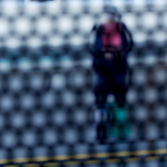 Urban Commute - Series No.2: 9153 - Close Detail View, fine art photograph by Raymond Mather showing a blurred figure in red and navy with a backpack and helmet, standing before a row of colourful train seats. Seen through a circular grid, the image evokes themes of pause, movement, and visual abstraction.