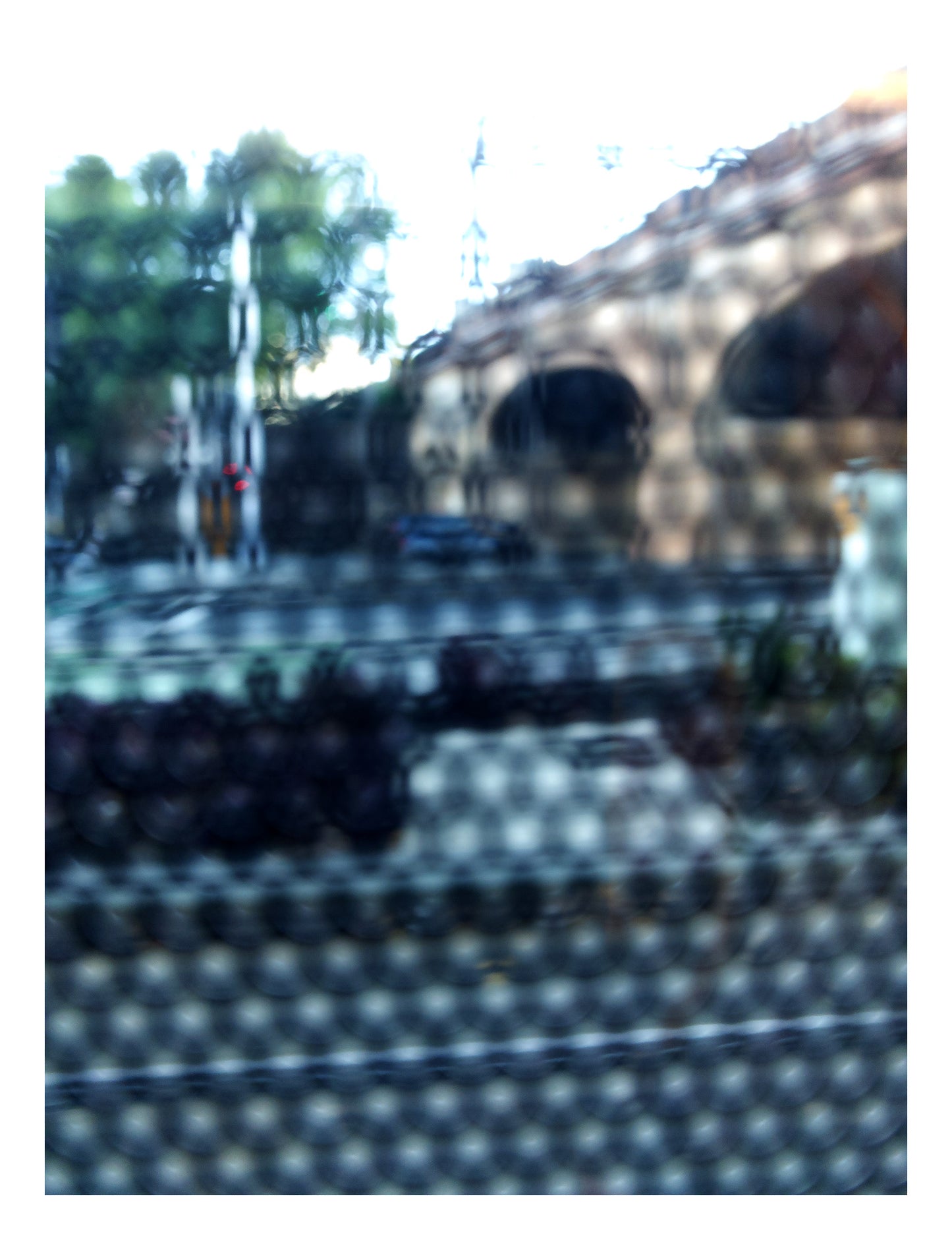 Urban Commute - Series No.2: 9164v1 - 81.5x104cm, fine art photograph by Raymond Mather featuring a city street scene with a brick railway viaduct, trees, and soft reflections, viewed through a patterned surface. The image is blurred and dappled, evoking a quiet, abstracted sense of place and structure.