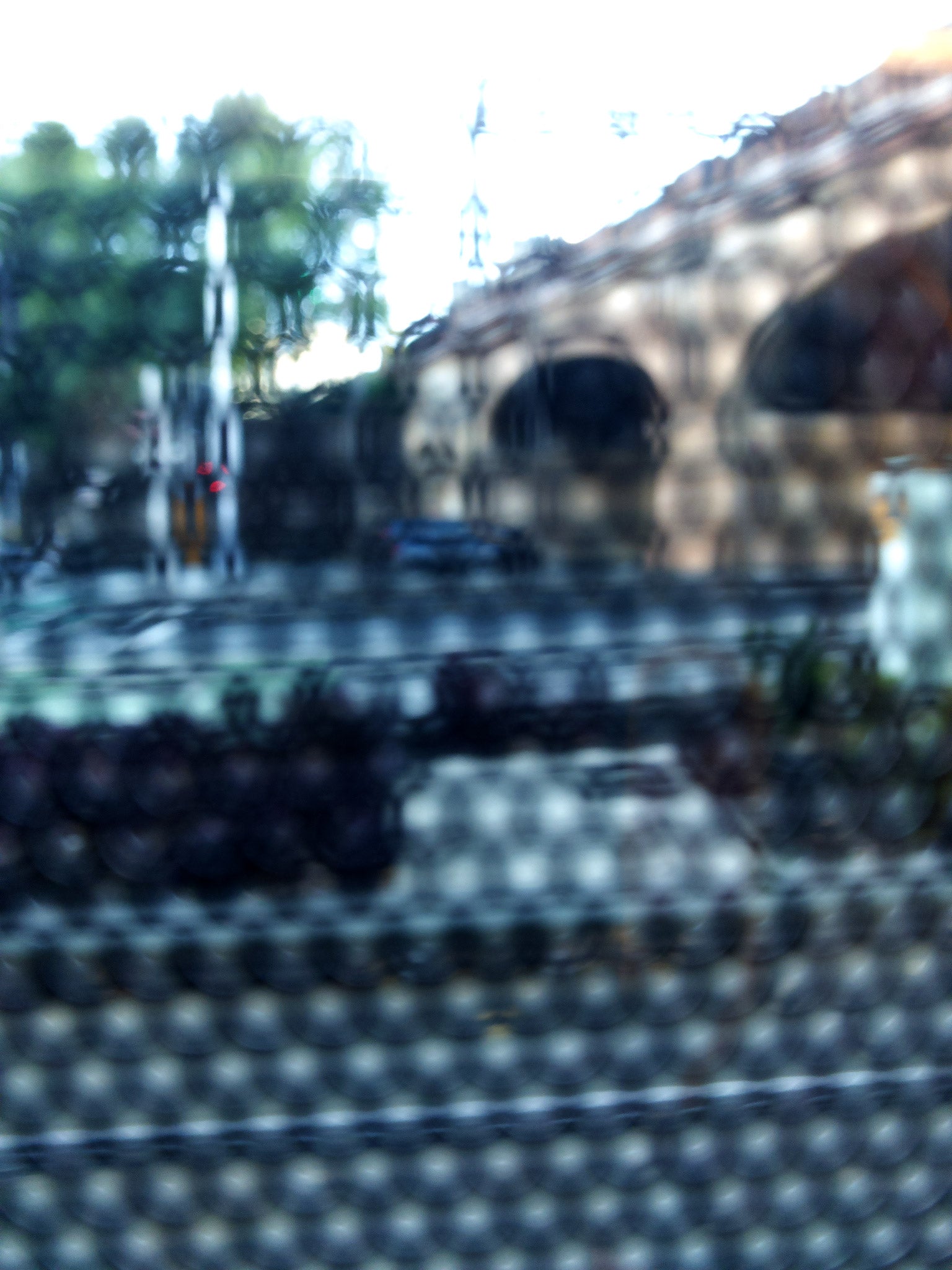 Urban Commute - Series No.2: 9164v1 - 67.5x90cm, fine art photograph by Raymond Mather featuring a city street scene with a brick railway viaduct, trees, and soft reflections, viewed through a patterned surface. The image is blurred and dappled, evoking a quiet, abstracted sense of place and structure.