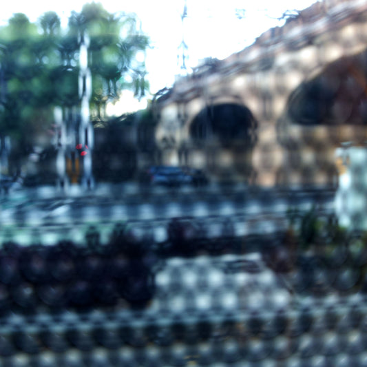 Urban Commute - Series No.2: 9164v1 - Close Detail View, fine art photograph by Raymond Mather featuring a city street scene with a brick railway viaduct, trees, and soft reflections, viewed through a patterned surface. The image is blurred and dappled, evoking a quiet, abstracted sense of place and structure.