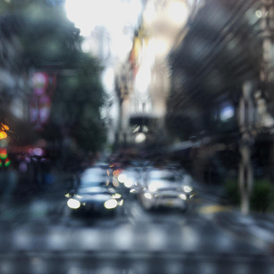Urban Commute - Series No.2: 9201(1)v1a - Close Detail View, fine art photograph by Raymond Mather showing blurred cars with headlights approaching an intersection, viewed through a patterned surface. City buildings rise on either side, creating a dreamlike abstract of light, rhythm, and perspective.